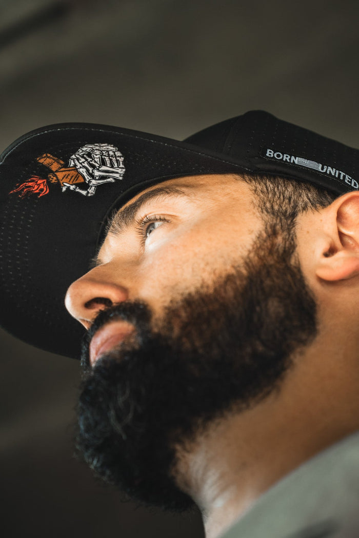A bearded man wearing the Born United Premium Snapback - Burning Bridges by Born United Private Label, featuring embroidered designs and “BORN UNITED” on the side, looks upward. Subtle "Burning Bridges" details accent the black cap. Shot from a low angle in soft light.