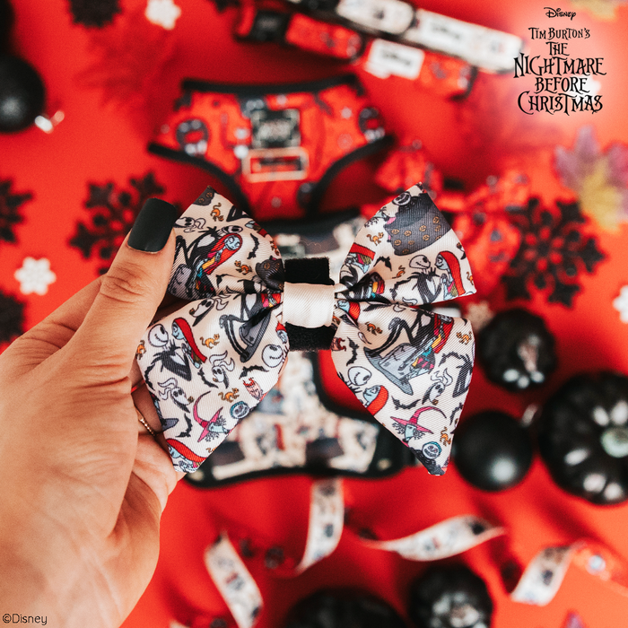 A hand holds the Sassy Woof Dog Sailor Bow - The Nightmare Before Christmas™ (Beige) against a red backdrop with themed ornaments—perfect for pairing with your dog’s collar or harness if you’re a fan of the movie.