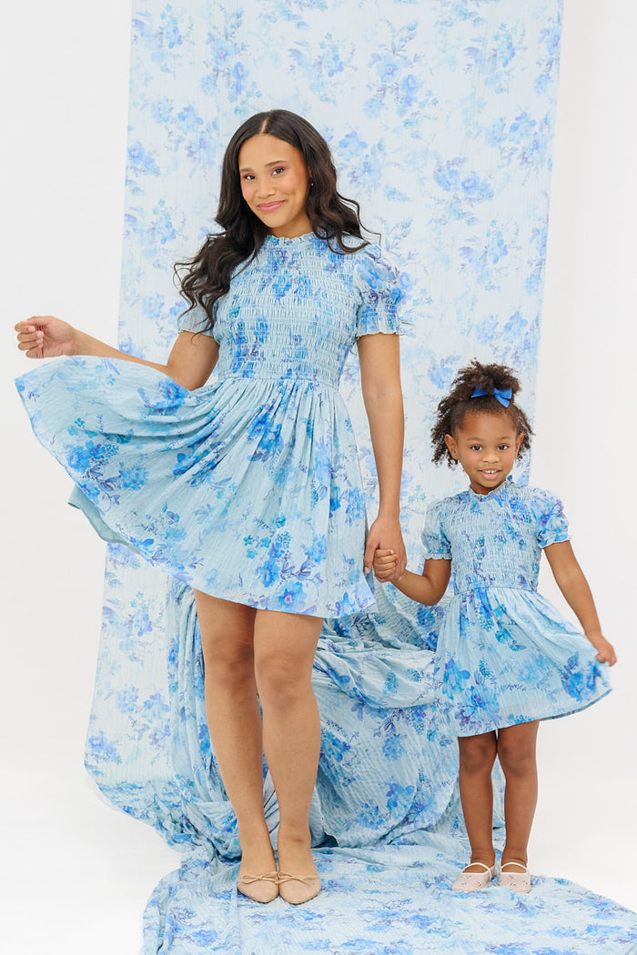 A woman and a young girl smile and hold hands in Ivy City Co’s Mini Flynn Dress in Aquarelle Fleur, both wearing matching blue watercolor floral outfits. The coordinated backdrop highlights their charming spring style.