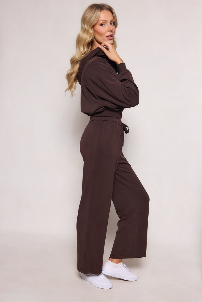 A woman with long blonde hair wears Ivy City Co's Amara Sweatpant in Espresso, featuring a relaxed leg and elastic waistband, paired with white sneakers. She stands in profile, hand to chin, looking at the camera against a plain light background.