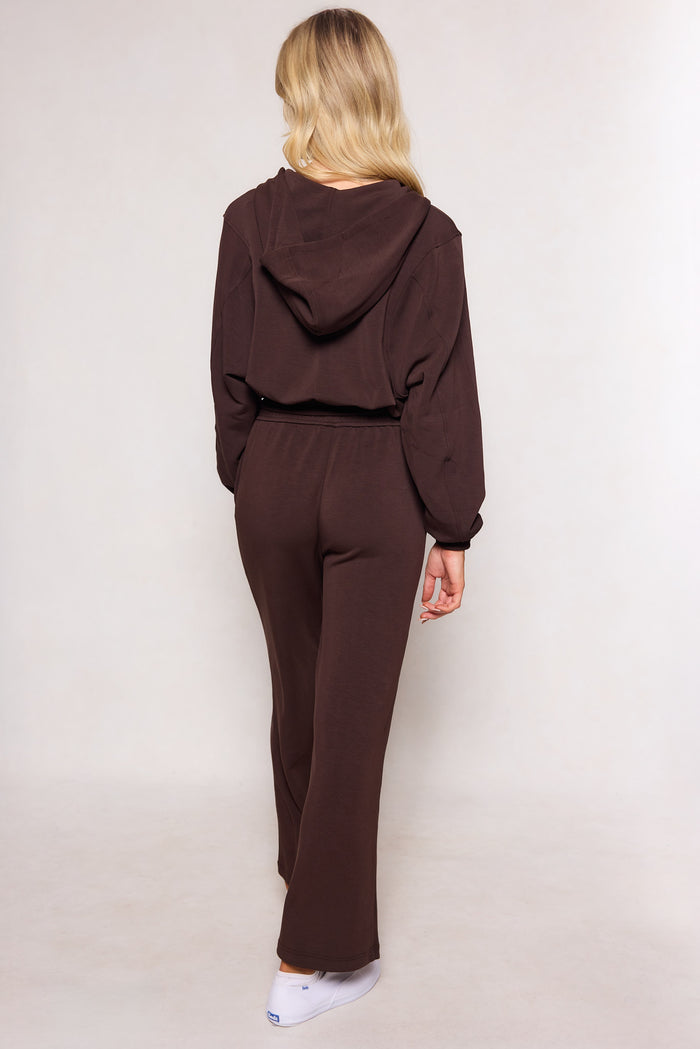 A woman with long blonde hair models the Ivy City Co Amara Sweatpant in Espresso, featuring a relaxed leg and elastic waistband, as she stands facing away from the camera against a plain light background.