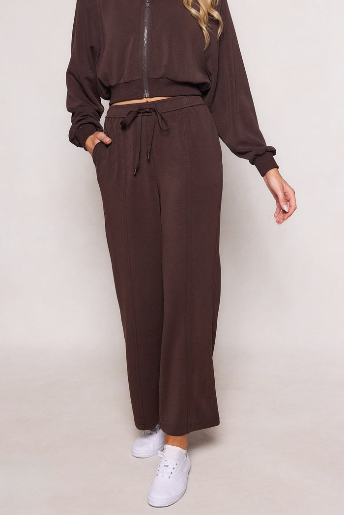A person wearing the Ivy City Co Amara Sweatpant in Espresso, featuring an elastic waistband and relaxed leg, paired with a brown zip-up jacket and white sneakers. The individual stands with one hand in their pocket, and their head is not visible.