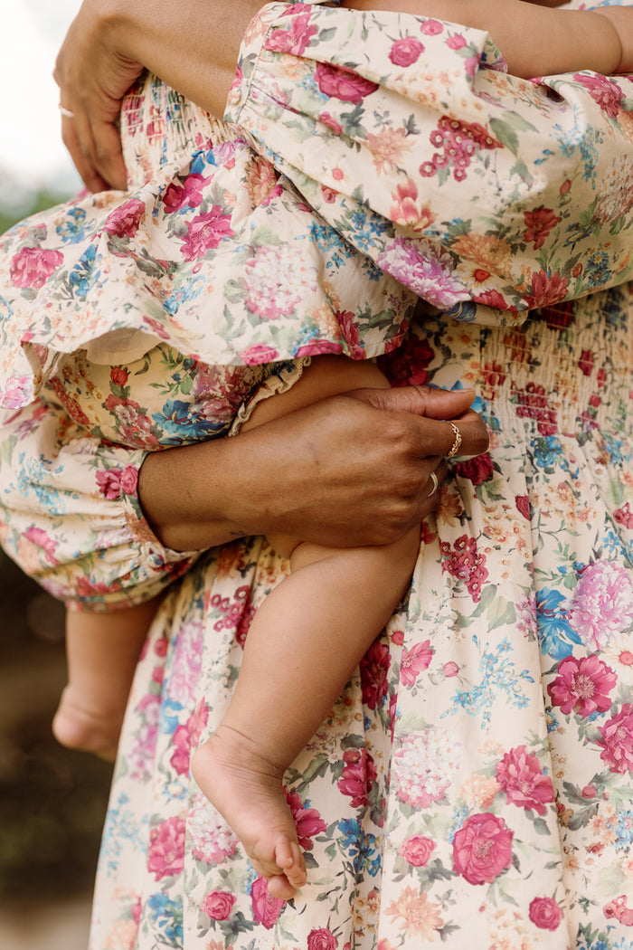 A close-up shot of a person with medium-toned skin wearing a floral dress, lovingly cradling a baby who is also dressed in the Ivy City Co Baby Adelpha Dress Set in Autumn Amity Floral. The focus is on the parent’s hand holding the baby’s leg, highlighting the delicate smocked cotton dress that perfectly complements their coordinated style.