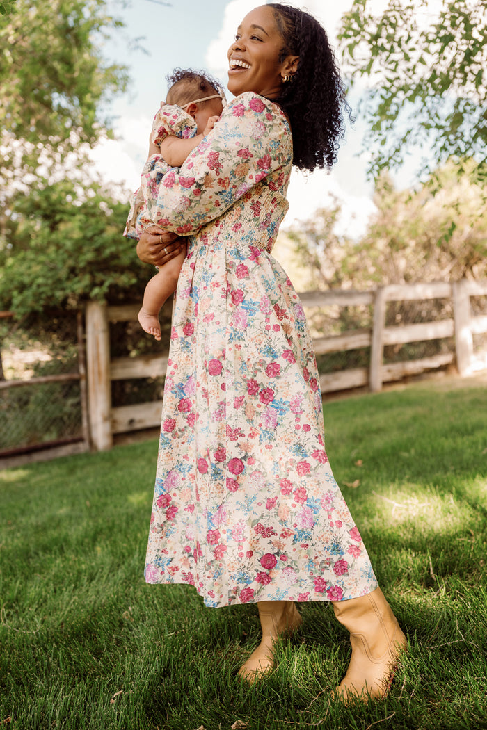 A woman stands on the grass, smiling and holding a baby dressed in the Ivy City Co Baby Adelpha Dress Set in Autumn Amity Floral; both wear smocked cotton dresses, with trees and a wooden fence in the background.