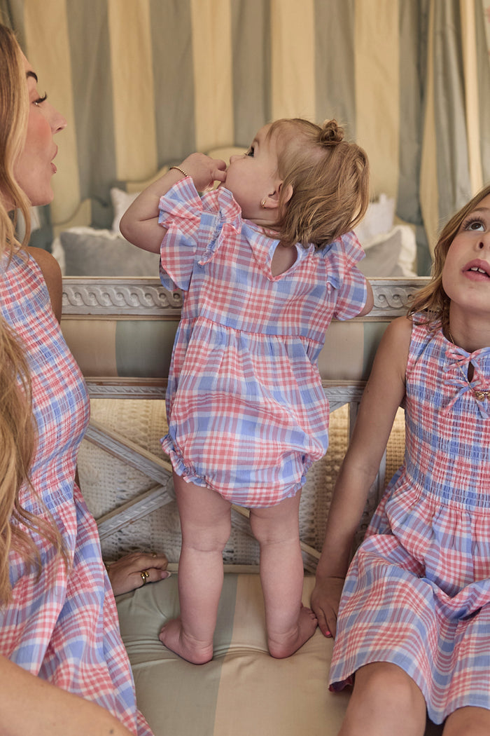 A baby in the Ivy City Co Baby Ana Romper in Blossom Plaid stands on a couch between two girls in matching dresses. One girl sits on the right looking up, while a woman is partially seen on the left among cushions and draped fabric.