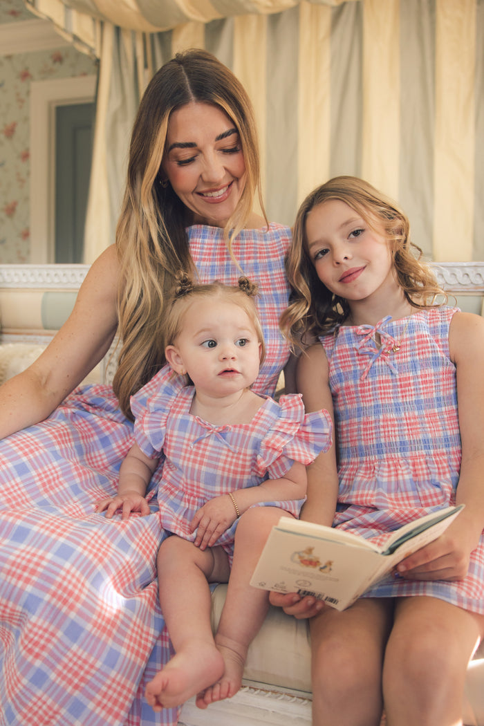 A woman and two young girls sit together on a sofa, all wearing the Ivy City Co Baby Ana Romper in Blossom Plaid. The older girl holds an open book while the woman smiles, creating a cozy spring vibe in this softly lit scene.