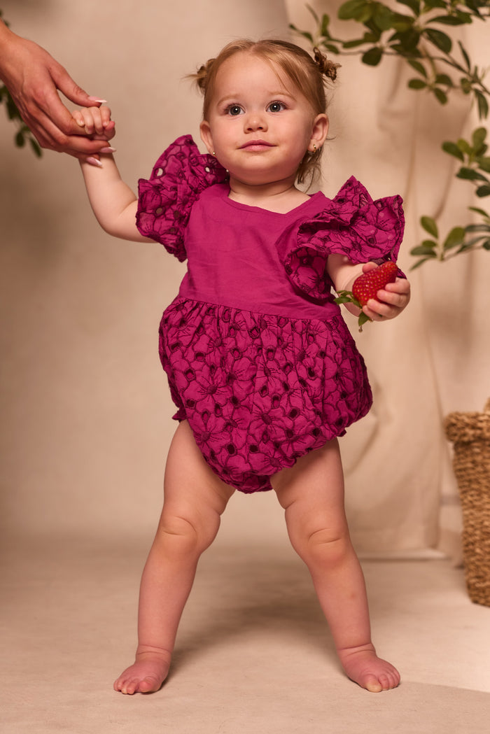 A toddler wearing the Ivy City Co Baby Hattie Romper in Magenta holds a strawberry and an adult’s hand, standing barefoot indoors near a potted plant.
