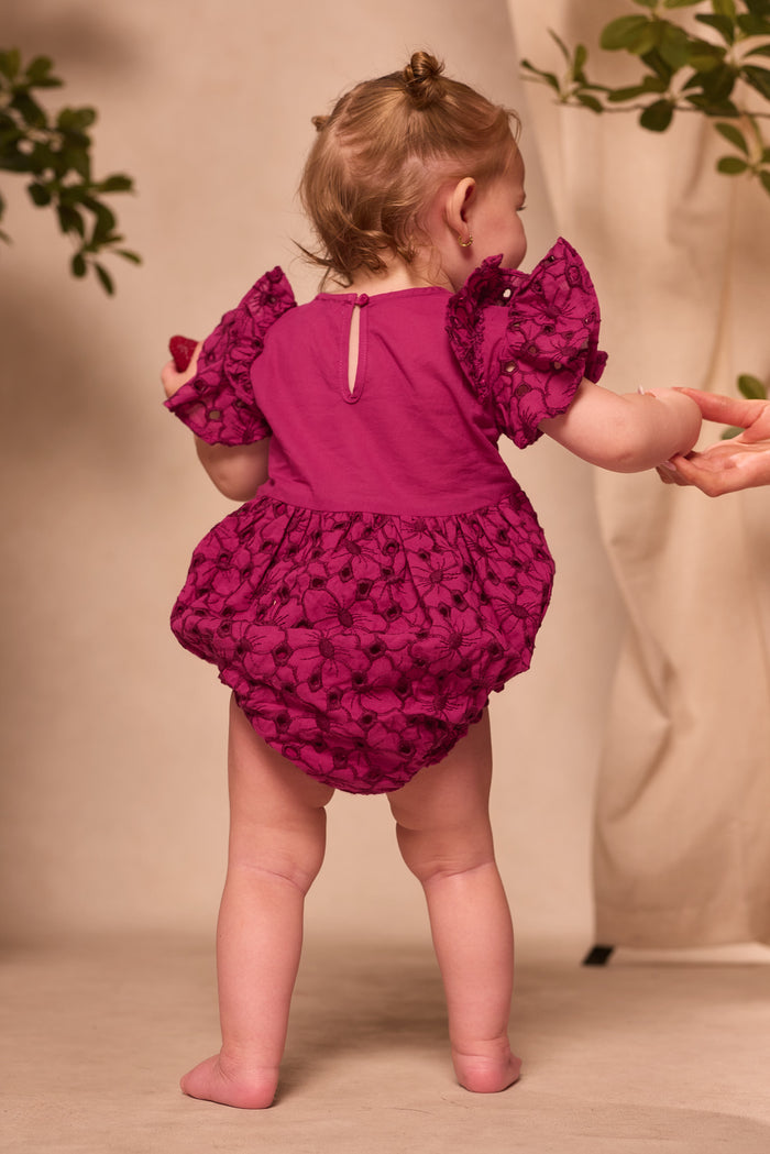 A baby stands barefoot, facing away, in the Ivy City Co Baby Hattie Romper in Magenta with puffed sleeves. An adult hand gently holds the baby's arm, while greenery appears in the background.