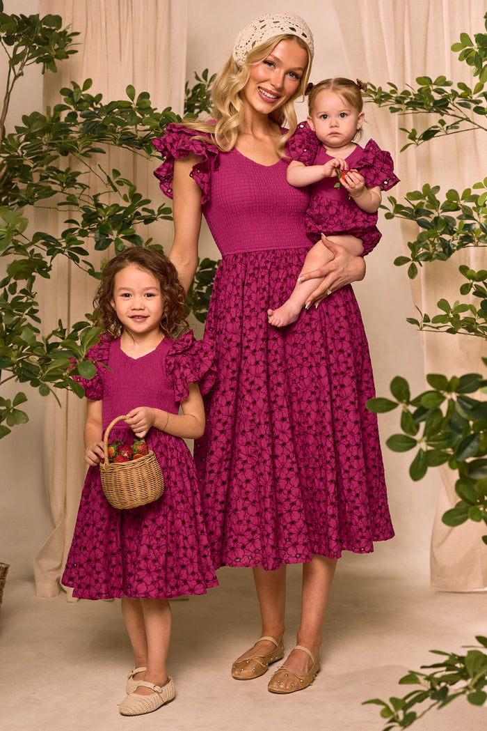 A woman and two girls wear matching magenta eyelet cotton outfits with ruffled sleeves. The baby shines in the Ivy City Co Baby Hattie Romper in Magenta—an adorable choice for Easter among greenery and beige curtains.
