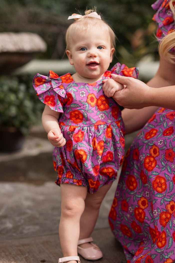 A baby stands outdoors holding an adult’s hand, wearing the Ivy City Co Baby Hattie Romper in Thistle Flower Poppy Woodblock. The adult is in a matching floral dress, with greenery softly blurred in the background.