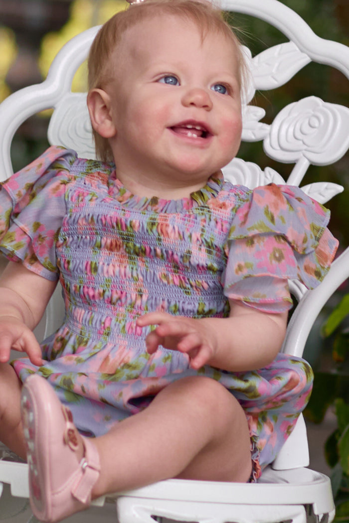 A smiling toddler with light hair sits on a white ornate chair wearing the Ivy City Co Baby Lottie Romper in Vibrant English Garden, paired with pale pink shoes and surrounded by greenery—perfect for a garden-inspired look.