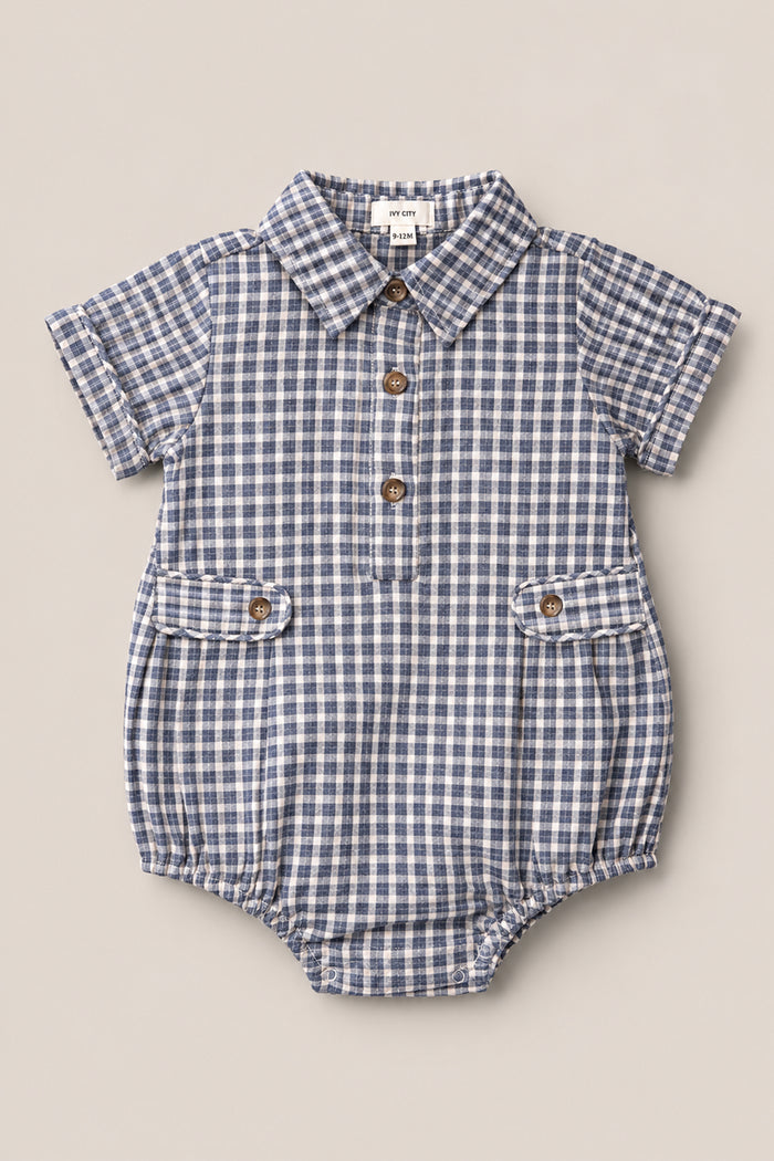 The Baby Lucas Romper in Prussian Blue Plaid by Ivy City Co features a classic collar neckline, short sleeves, three front buttons, and two front pockets with button closures. Crafted from cotton, this blue and white plaid romper combines timeless style and comfort for your little one.