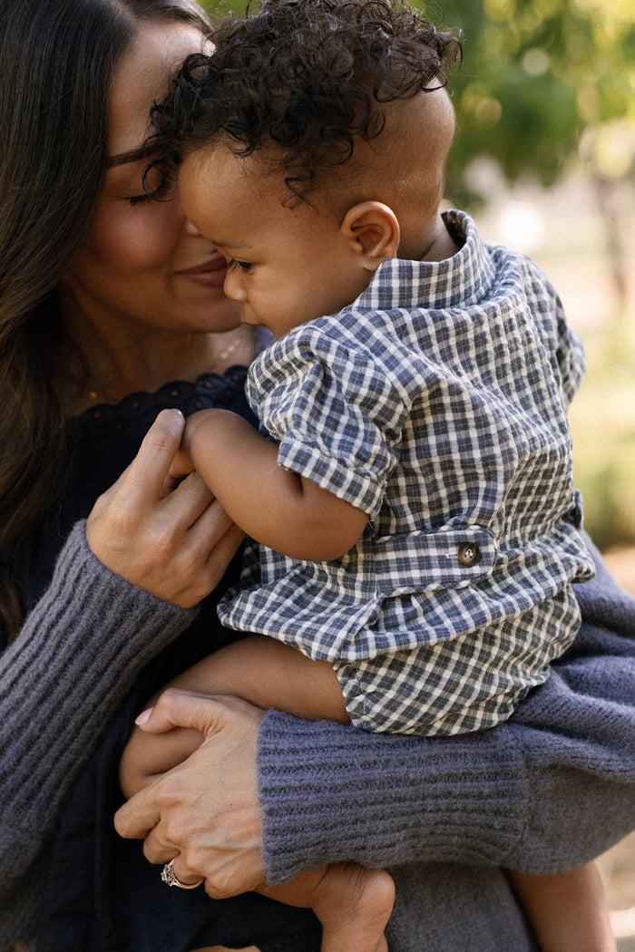 A woman in a gray sweater lovingly holds a smiling baby wearing the Ivy City Co Baby Lucas Romper in Prussian Blue Plaid, featuring a charming collar neckline, their faces gently touching outdoors.