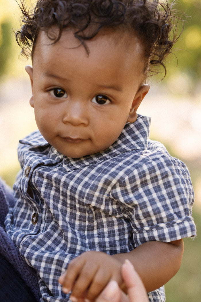A baby with curly hair wearing the Ivy City Co Baby Lucas Romper in Prussian Blue Plaid, featuring a collared neckline, looks at the camera while being held outdoors.
