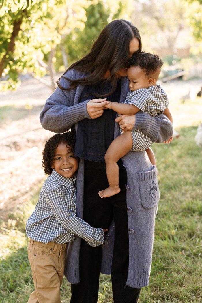 A woman in a purple cardigan holds a small child dressed in Ivy City Co’s Baby Lucas Romper in Prussian Blue Plaid while another child hugs her leg. They are outside on a grassy area with trees in the background, all smiling and relaxed together.