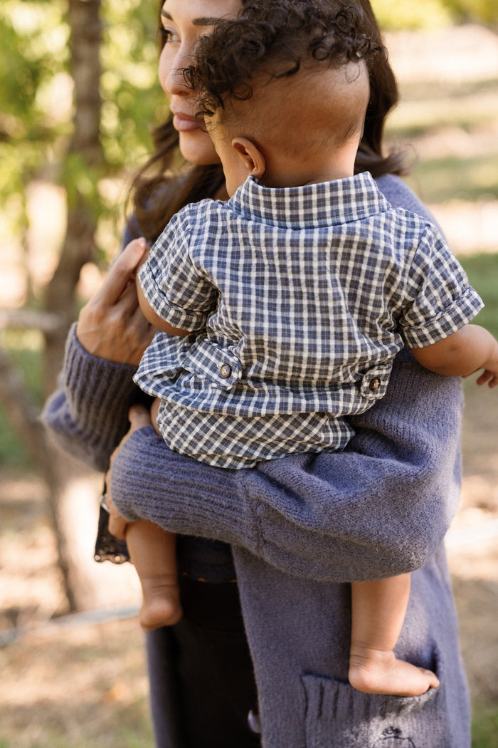 A woman holds a baby outdoors, who is dressed in the Ivy City Co Baby Lucas Romper in Prussian Blue Plaid, featuring a cotton fabric and a collared neckline. The baby faces away while the woman, partially visible in a blue sweater, stands amid sunlit trees.