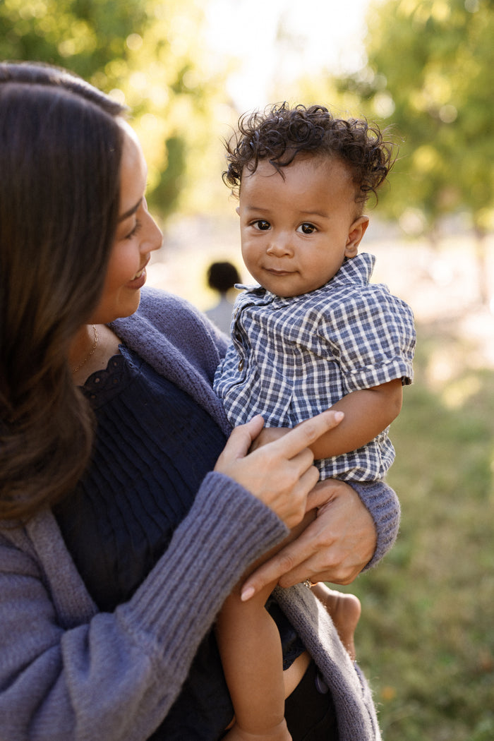 A woman holds a baby outdoors, both looking content. The baby is wearing the Ivy City Co Baby Lucas Romper in Prussian Blue Plaid, featuring a collared neckline. Greenery and soft sunlight fill the background.