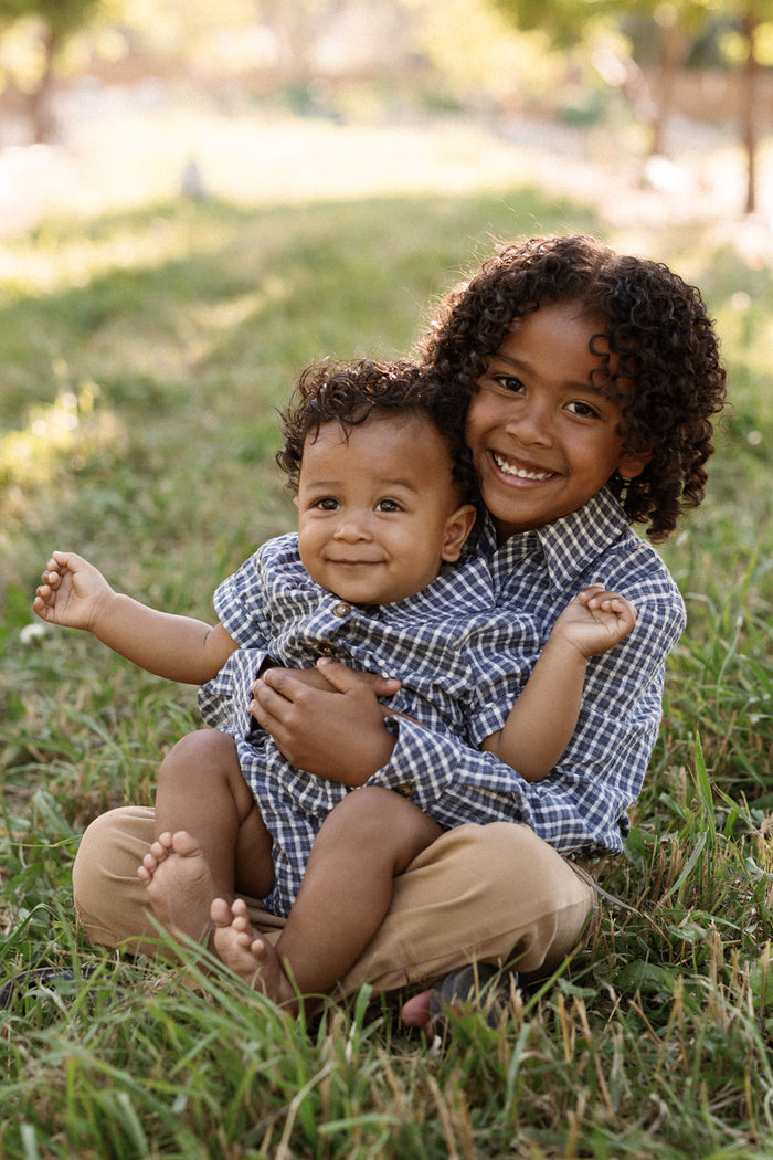 Two young children sit together on the grass outdoors, smiling warmly. The older child hugs the younger from behind as both wear matching Ivy City Co Baby Lucas Romper in Prussian Blue Plaid, featuring a smart collared neckline. Sunlight creates a gentle, golden glow in the background.