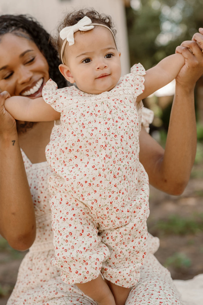 A smiling woman holds up a baby dressed in the Ivy City Co Baby Mirabelle Romper in Cream Ditsy Floral with a white bow headband. They are outdoors, and the baby’s arms are stretched outward.