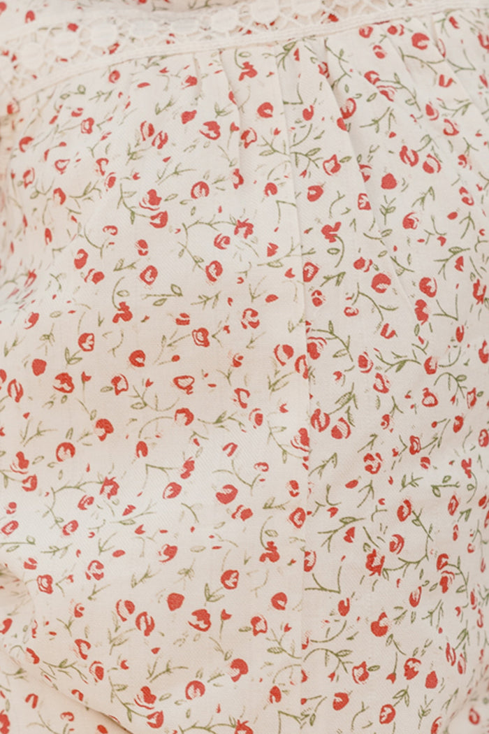 Close-up of the Ivy City Co Baby Mirabelle Romper in Cream Ditsy Floral, showcasing its cream-colored cotton fabric adorned with a delicate pattern of small red flowers and green stems, highlighted by gentle gathering and a lace trim along the top edge.
