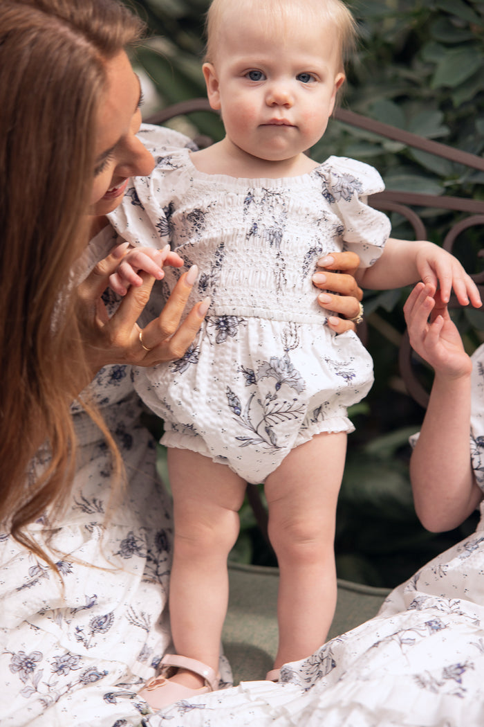 A baby with light hair wears the Ivy City Co Baby Roselyn Romper in Blue Enchanted Floral, standing while an adult with long brown hair gently holds them. Both are dressed in matching blue floral outfits, surrounded by greenery.