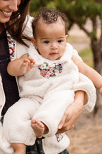 An adult holds a baby outdoors, dressed in the Ivy City Co Baby Siena Onesie in Ivory & Midnight Garden—a soft sherpa piece featuring a delicate floral pattern. The baby, with curly hair and an arm raised, sits securely while only part of the adult’s face is visible.