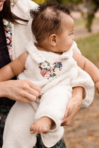 An infant wearing the Ivy City Co Baby Siena Onesie in Ivory & Midnight Garden sits on an adult’s lap outdoors. The little one, dressed in the cozy ivory sherpa outfit with a floral heart patch, gazes to the side as the adult gently supports their arm and leg.