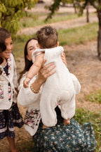 A woman in a green dress lifts a baby dressed in the Ivy City Co Baby Siena Onesie in Ivory & Midnight Garden outdoors, while a smiling young girl stands nearby, surrounded by grass and trees.