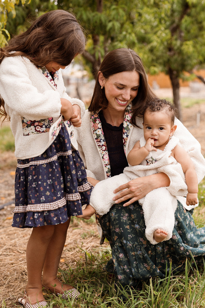 A woman kneels on the grass, smiling at a baby in her lap dressed in the Ivy City Co Baby Siena Onesie in Ivory & Midnight Garden. A young girl stands beside them, looking down. All three wear floral-patterned outfits and light jackets, with trees and greenery in the background.