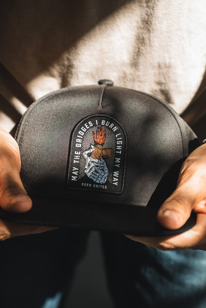 A person holds the Born United Premium Snapback - Burning Bridges by Born United Private Label, featuring a flaming hand patch and the quote "May the bridges I burn light my way," highlighted in sunlight to symbolize bold new paths.