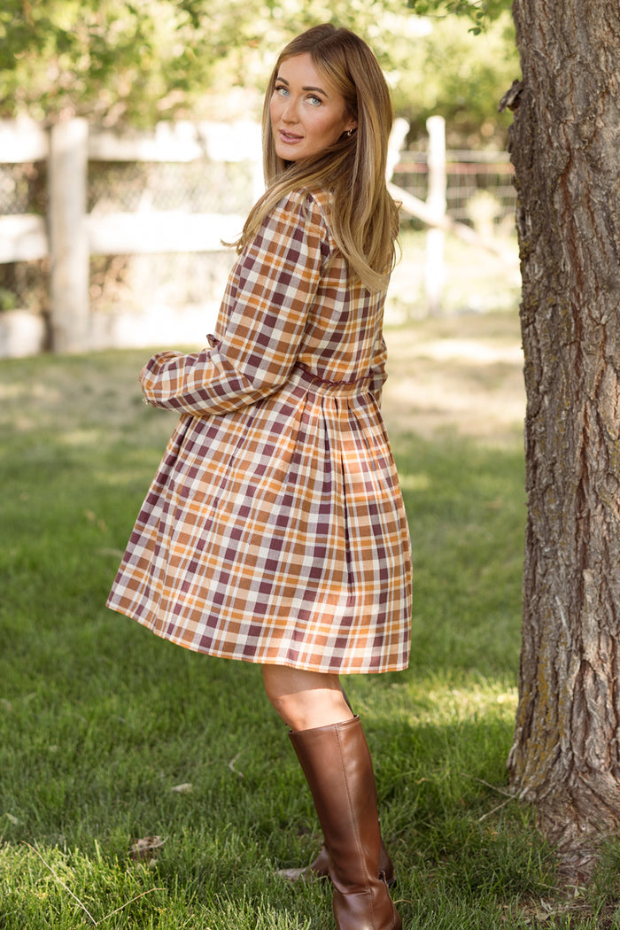 Wearing the Ivy City Co Bowie Long Sleeve Dress in Spiced Plaid and tall brown boots, a woman stands on grass, glancing over her shoulder with trees and a wooden fence in the background.