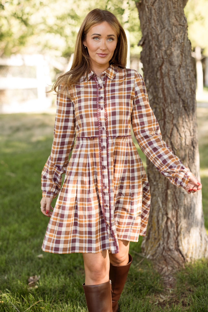 A woman wearing the Ivy City Co Bowie Long Sleeve Dress in Spiced Plaid with balloon sleeves and brown boots stands outside on green grass by a tree, looking at the camera while holding her dress with one hand.