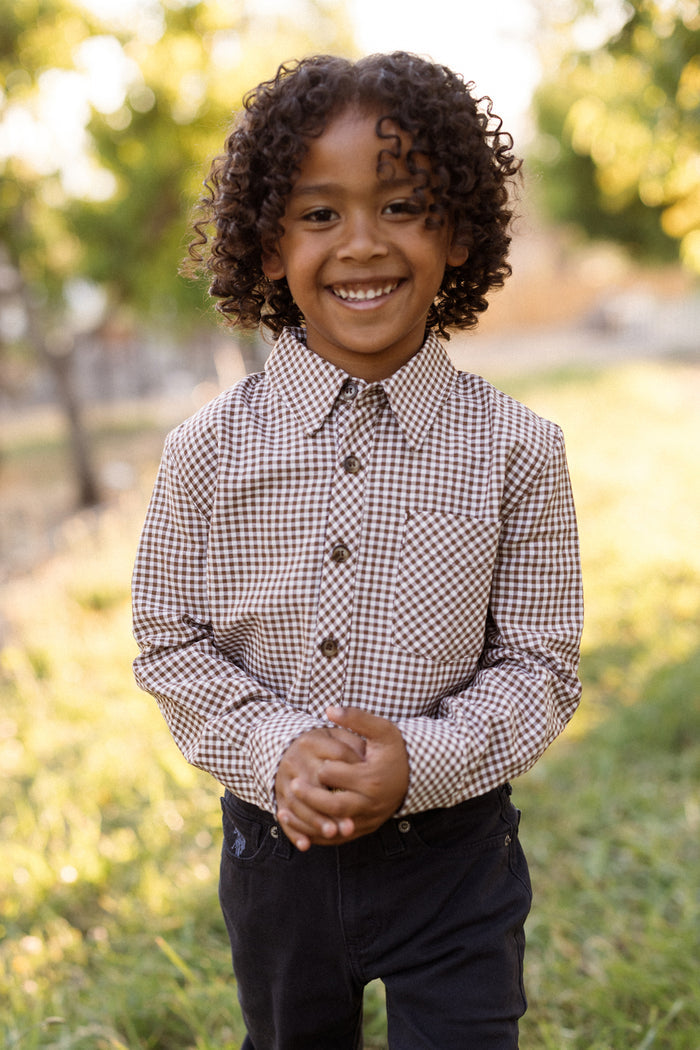 A smiling child with curly hair stands outside on the grass, wearing the Ivy City Co Boys John Shirt in Brown Gingham—a classic long-sleeve boys’ shirt made from soft woven cotton with a checkered pattern—paired with dark pants, and blurry trees in the background.