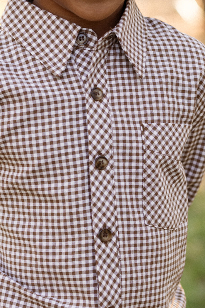 A close-up of a child’s torso wearing the Boys John Shirt in Brown Gingham by Ivy City Co, a woven cotton long sleeve boys shirt with a front pocket. The background is blurred with green tones.
