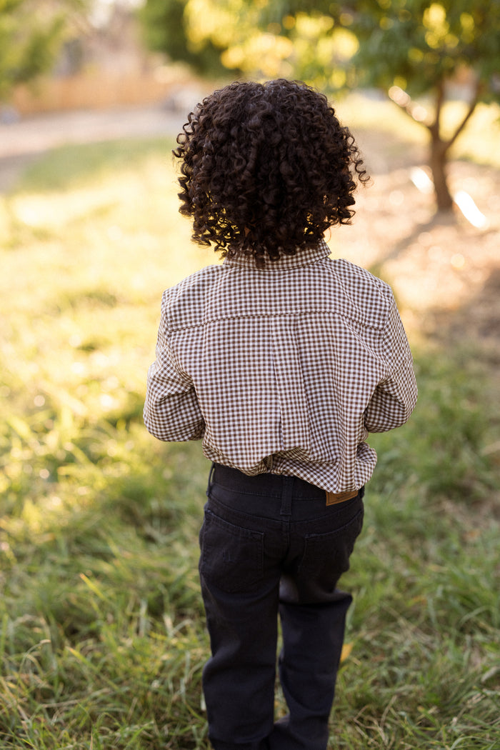 A young child with curly hair, wearing the Ivy City Co Boys John Shirt in Brown Gingham and dark pants, stands outside on the grass facing away from the camera toward sunlight and trees.