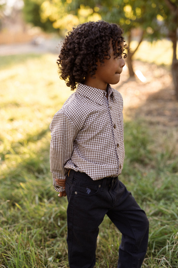 A young child with curly hair stands sideways outdoors in a sunlit grassy area, wearing the Ivy City Co Boys John Shirt in Brown Gingham and dark pants, with hands behind their back.