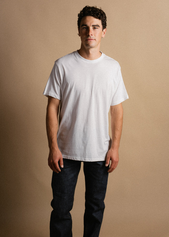 Man in a white T-shirt and dark jeans stands against a beige background, looking confidently at the camera.