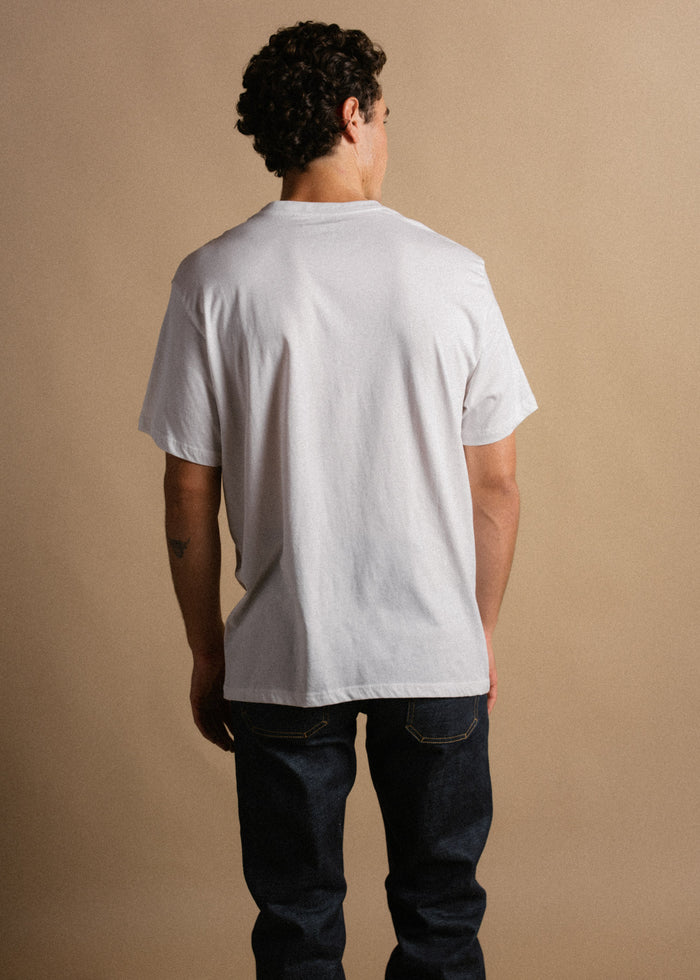 Man in white t-shirt and jeans stands with back to camera against beige background, showcasing casual fashion style.