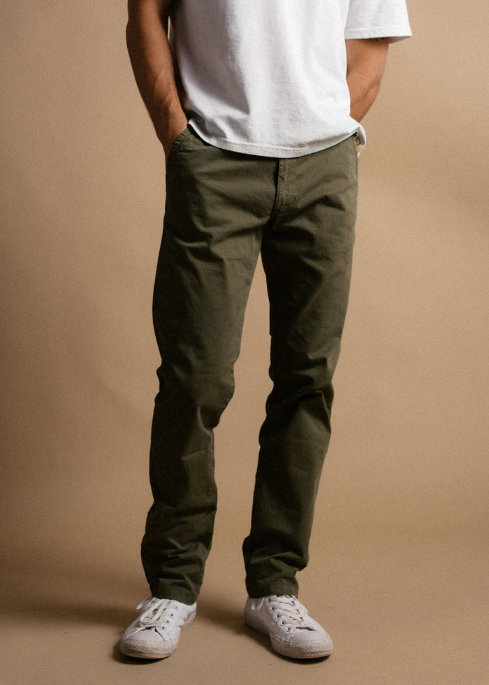 Man in a white t-shirt, olive green pants, and white sneakers stands against a beige background with hands in pockets.