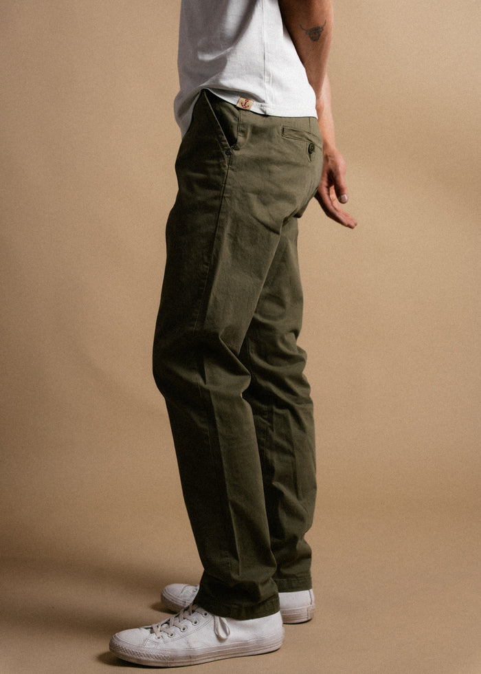 Man in olive green pants and white sneakers stands sideways against a beige background, wearing a white shirt with a tattoo visible.