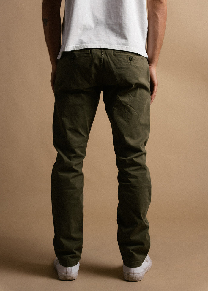Man in olive green pants and white sneakers stands against a beige background, wearing a white shirt, viewed from the back.