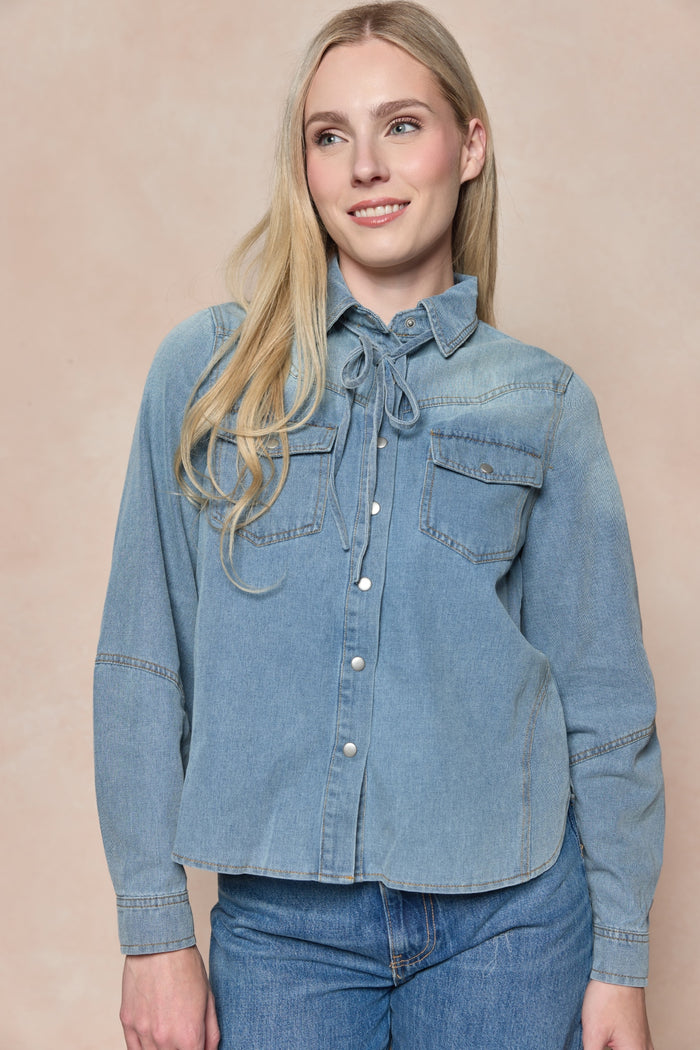 A woman with long blonde hair wears the Ivy City Co Denver Top in Lightwash Denim, featuring chest pockets, paired with blue jeans. She stands against a beige background, smiling gently and glancing to the side.