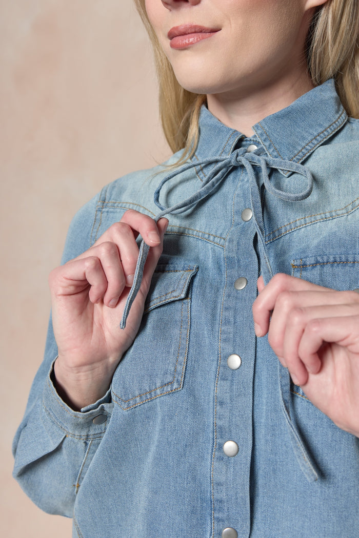 A woman ties the thin denim strings at the collar of the Ivy City Co Denver Top in Lightwash Denim. Only her lower face and torso are shown. The blouse features pearl snap buttons and classic chest pockets.