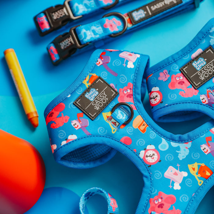 Sassy Woof’s Dog Adjustable Harness - Blue's Clues™ features vibrant cartoon prints and is displayed with matching leashes and accessories on a blue and orange backdrop, accented by a yellow crayon and an orange toy.