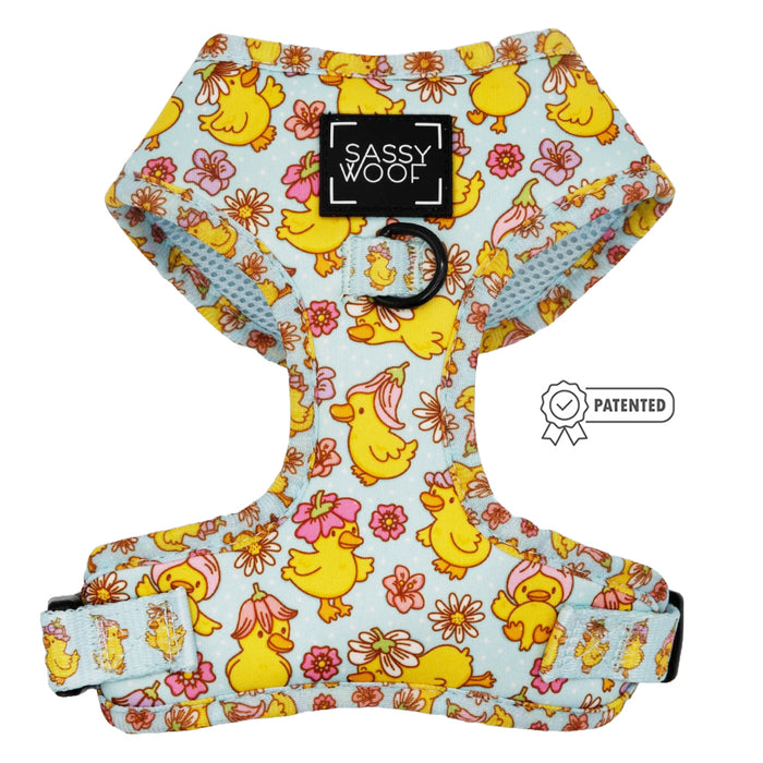 The Sassy Woof Dog Adjustable Harness - Good Duck, Babe! is a light blue harness with yellow cartoon ducks and pink flowers. It features a Sassy Woof label, metal leash ring, and an adjustable strap with patented design badge.