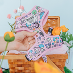 A hand displays the Sassy Woof Dog Adjustable Harness - Hello Kitty® & Friends Flower Festival, a pastel pink harness with Sanrio characters and doodles, set against a wicker picnic basket and flowers.