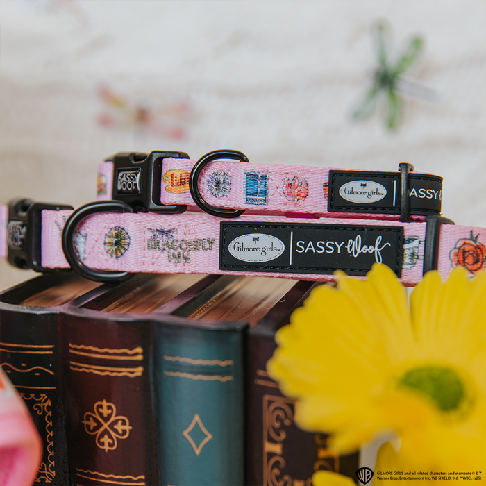 Two pink Dog Collar - Gilmore Girls™ by Sassy Woof from the Stars Hollow pet collection sit atop stacked ornate books, with a yellow flower in front and blurred butterfly decor in the background.