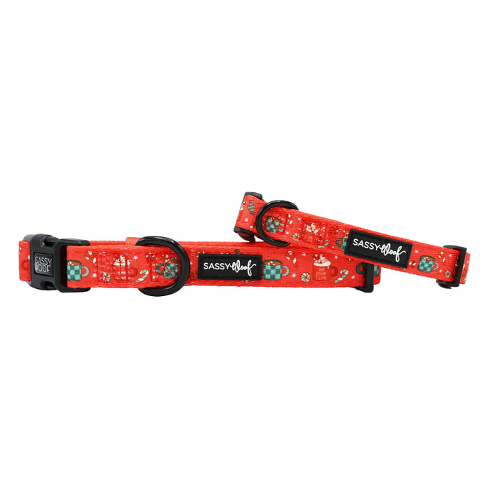 Two Sassy Woof Dog Collars from the Hot Cocoa Club collection with festive holiday patterns—paw prints, bones, and snowflakes—each with a black plastic buckle and Sassy Woof label. Both are adjustable.
