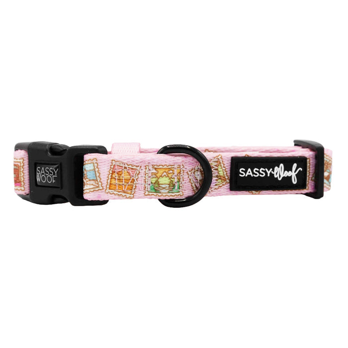 The Sassy Woof Dog Collar - Out of Office features a pink, adjustable design with colorful postage stamps, a black buckle, and logo tag. It's part of the Puppy Growth Pawtection Plan for growing pups.