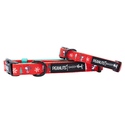 Two red Sassy Woof dog collars featuring festive Peanuts™ Beagle Bells designs and black buckles—each labeled "PEANUTS" and "SASSY WOOF." Perfect holiday accessories for your pup!.